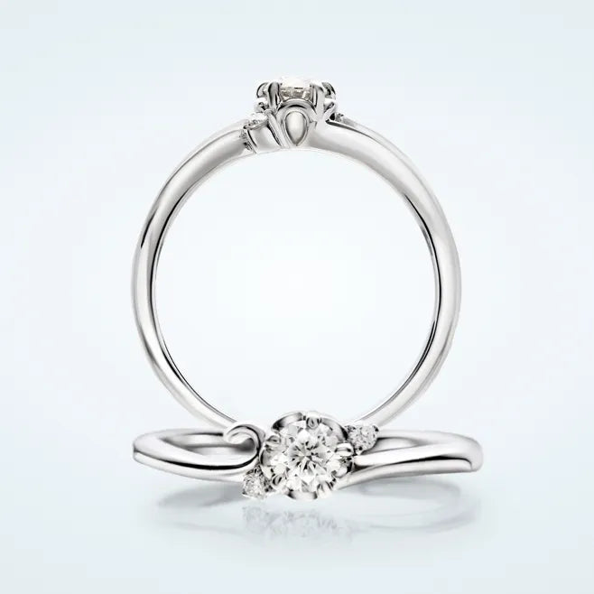 花びらデザインのリング枠に、センターの 0.2ct 天然ダイヤモンドを 5本爪で留めたエンゲージリングです。センターに寄りそうように、両サイドへメレダイヤモンド 2石（計0.03ct）を添えています。